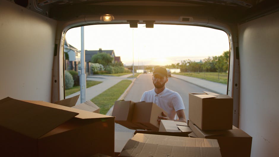 A smiling man with a full beard and curly black hair stands outdoors next to a white moving van, which displays the words 'MOVING COMPANY' and 'LOCAL & LONG DISTANCE' in bold black letters. He is dres