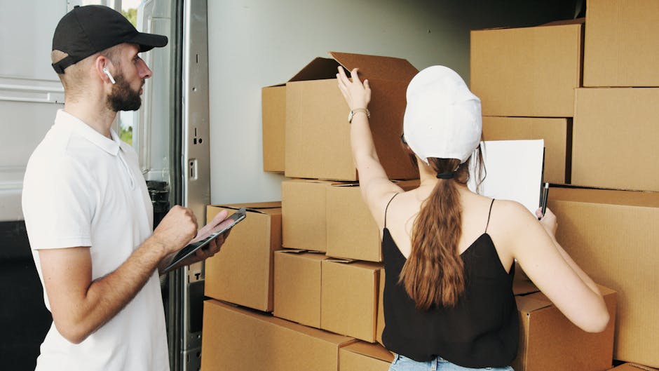 A man and woman engaged in the home relocation process next to a large open van. The man, wearing a white t-shirt, a black cap, and wireless earbuds, is holding a clipboard or tablet and observing. The woman, dressed in a black sleeveless top and a white cap, is standing with her back to the camera, reaching up to adjust or examine a cardboard box on top of a stack inside the van. The boxes, made of corrugated cardboard, are arranged in multiple rows and columns, with some open and others sealed. The scene takes place near the entrance of a property, with natural light illuminating the area, indicating daytime. The van, which is likely used by Man With a Van Lisson Grove, is stationed on a paved driveway or pavement, actively involved in furniture transport and packing and moving operations as part of a professional removals service in Lisson Grove.