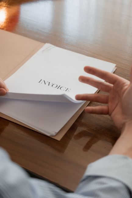 Close-up of a person’s hand reaching towards a white invoice document placed on top of a cardboard folder, sitting on a wooden surface. The invoice has the word 'INVOICE' printed prominently at the to