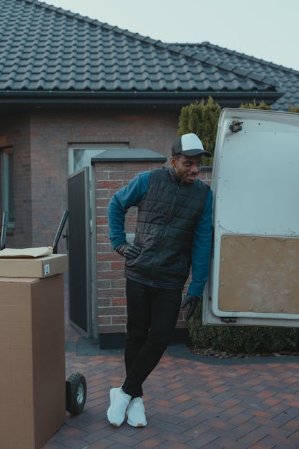 A man wearing a grey cap, blue long-sleeve shirt, black padded vest, and black pants, stands beside a white moving van with an open side door, parked on a paved driveway in front of a residential property with a brick exterior and a tiled roof. He is leaning against the van with one hand on his hip and appears to be resting during the home relocation process. Nearby, there are cardboard boxes, some with packing tape, placed on a dolly with black wheels. The scene suggests a furniture transport or packing and moving activity as part of a house removals service, with the man preparing or taking a break during the loading or unloading process, which is common in professional removals like those offered by Man With a Van Lisson Grove.