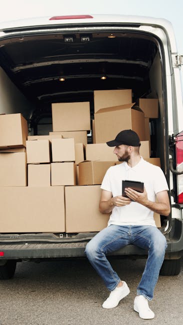 A man wearing a black cap, white t-shirt, and blue jeans is seated on the pavement in front of a large, open van used for home relocation. The van's interior is filled with numerous cardboard boxes of various sizes, some stacked on top of each other and others placed on the floor, along with packaging materials such as plastic wraps and packing paper. The man is holding a tablet device and appears to be reviewing or organizing the packing process. The scene shows the loading process involved in furniture transport and packing and moving services, with the van positioned on a street or driveway, ready for further transport, while [COMPANY_NAME], Man With a Van Lisson Grove, specializes in removal and house moving services in the Lisson Grove area.