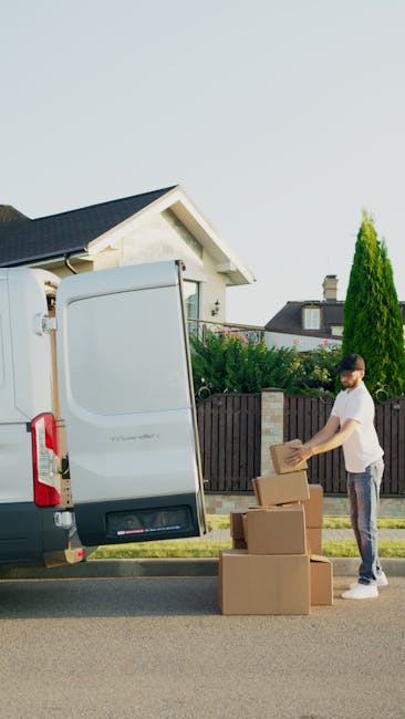 A man wearing a grey cap, blue long-sleeve shirt, black padded vest, and black pants, stands beside a white moving van with an open side door, parked on a paved driveway in front of a residential property with a brick exterior and a tiled roof. He is leaning against the van with one hand on his hip and appears to be resting during the home relocation process. Nearby, there are cardboard boxes, some with packing tape, placed on a dolly with black wheels. The scene suggests a furniture transport or packing and moving activity as part of a house removals service, with the man preparing or taking a break during the loading or unloading process, which is common in professional removals like those offered by Man With a Van Lisson Grove.