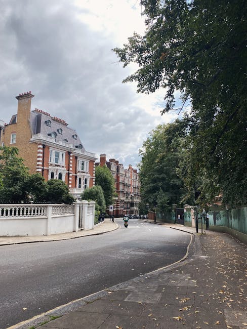 A quiet street scene featuring a row of historic red-brick residential buildings with ornate architectural details, situated adjacent to a pavement lined with mature trees providing partial shade. The street has a gentle curve and appears to be part of a residential neighbourhood in Lisson Grove. The sky is overcast with grey clouds, and the street is empty of cars and pedestrians. This image reflects a typical environment where house removals or furniture transport from a property on Harley Street may be planned, with surrounding greenery and urban architecture indicating a potentially accessible location for moving services by Man With a Van Lisson Grove.