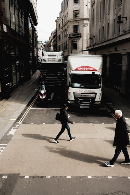 A narrow street scene showing two delivery vans parked parallel to each other, with the one on the right branded 'Uniserve' and the one behind labeled 'Becton Transport.' The vans are positioned close to the curb, partially blocking the roadway. In front of the vans, a black scooter is parked on a dedicated loading zone with railway track markings visible on the pavement. Two pedestrians are crossing the street: an older man with white hair wearing a dark coat and walking on the right, and a younger woman carrying a backpack and wearing casual clothing, walking towards the left. The street is lined with tall, multi-storey buildings with classic architectural features, including fire escapes and balconies, under an overcast sky. The scene captures a typical urban environment suitable for house removals or moving services in Lisson Grove, with visible elements indicative of a home relocation transport process, overseen by Man With a Van Lisson Grove.