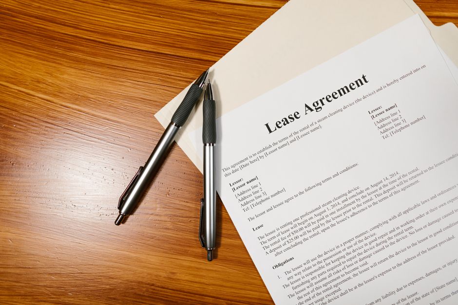 A lease agreement document printed on white paper placed on a wooden surface, with two black and silver ballpoint pens resting on top of it. The document features the title 'Lease Agreement' in bold at the top, followed by sections detailing the lease terms, landlord and tenant information, and obligations. The paper is partially angled, and the pens are positioned parallel to each other, with one pen slightly overlapping the other. The wooden surface has a warm tone with a smooth finish, and a corner of a light-colored folder or envelope is visible beneath the lease document. This image illustrates components involved in the administrative and planning aspects of house removals or home relocation processes, as handled by professionals such as Man With a Van Lisson Grove.
