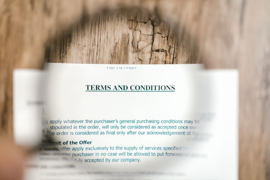 A close-up view of a printed document titled 'TERMS AND CONDITIONS' that is partially obscured by a blurred, circular focus in the foreground. The document is placed on a wooden surface inside a property, with the top edge slightly curling upward. The text on the paper includes references to the purchase and supply of services, indicating a formal agreement related to home relocation or removals. The background features a textured wooden floor or table, and the lighting appears natural, highlighting the paper’s surface. This image is relevant to moving and packing processes associated with house removals, as handled by a company like Man With a Van Lisson Grove.