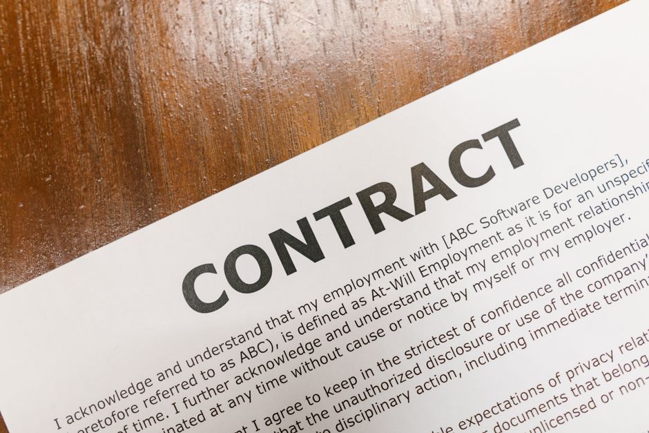 A lease agreement document printed on white paper placed on a wooden surface, with two black and silver ballpoint pens resting on top of it. The document features the title 'Lease Agreement' in bold at the top, followed by sections detailing the lease terms, landlord and tenant information, and obligations. The paper is partially angled, and the pens are positioned parallel to each other, with one pen slightly overlapping the other. The wooden surface has a warm tone with a smooth finish, and a corner of a light-colored folder or envelope is visible beneath the lease document. This image illustrates components involved in the administrative and planning aspects of house removals or home relocation processes, as handled by professionals such as Man With a Van Lisson Grove.