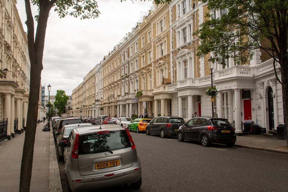 A residential street scene with rows of white terraced townhouses featuring ornate architectural details, including bay windows and decorative mouldings, on a cloudy day. Several parked cars of various models and colours line the curb, including a grey minivan in the foreground with the registration plate ND08 YBV, positioned close to the sidewalk. A green sports car and a black vehicle are visible further down the street. Trees with green foliage are situated along the pavement, providing shade and natural elements to the scene. The pavement is wide enough to accommodate loading and unloading activities, which are typical during home relocation or furniture transport processes. In the background, a hotel sign is visible on one of the buildings, indicating the mixed-use nature of the area. The overall environment reflects a typical London neighbourhood suitable for house removals managed by companies like Man With a Van Lisson Grove, with ample space for parking and moving equipment.
