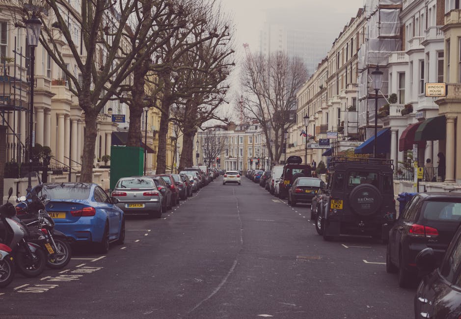 A quiet street scene in Lisson Grove during daytime with a row of parked cars lining both sides of the road, including sedans, hatchbacks, and an off-road vehicle, with some vehicles having visible registration plates. Several leafless trees are planted along the pavement, and historic white Victorian-style terraced houses with ornate facades and bay windows are visible on either side. A green public waste bin is positioned near the sidewalk on the left, and a few motorcycles are parked adjacent to the cars. The sky is overcast, and the street appears calm with minimal pedestrian activity. This setting may relate to home relocation or furniture transport processes, with no visible moving equipment or activity currently taking place. Man With a Van Lisson Grove occasionally operates in this area for removals and moving services, supported by parking tips for efficient loading and unloading near the homes.