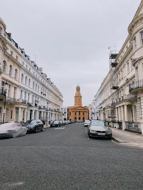 A residential street scene with rows of white terraced townhouses featuring ornate architectural details, including bay windows and decorative mouldings, on a cloudy day. Several parked cars of various models and colours line the curb, including a grey minivan in the foreground with the registration plate ND08 YBV, positioned close to the sidewalk. A green sports car and a black vehicle are visible further down the street. Trees with green foliage are situated along the pavement, providing shade and natural elements to the scene. The pavement is wide enough to accommodate loading and unloading activities, which are typical during home relocation or furniture transport processes. In the background, a hotel sign is visible on one of the buildings, indicating the mixed-use nature of the area. The overall environment reflects a typical London neighbourhood suitable for house removals managed by companies like Man With a Van Lisson Grove, with ample space for parking and moving equipment.
