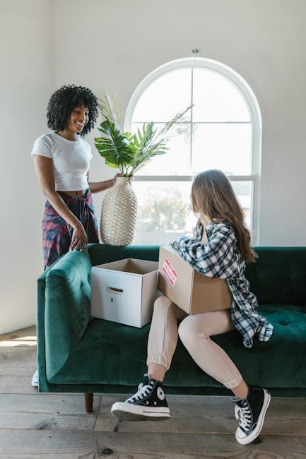 Inside a bright, minimally decorated room with a large arched window allowing natural light to fill the space, a woman with curly black hair, dressed in a white T-shirt and a plaid skirt, is standing beside a dark green upholstered sofa holding a large, white textured vase filled with green foliage. A young girl with long brown hair, wearing a plaid shirt, beige trousers, and black-and-white sneakers, is sitting on the sofa, holding a cardboard box with a red label and examining its contents. Next to her on the sofa are two open cardboard boxes, one of which is empty, while the other contains packing materials. The scene depicts a home relocation or packing process, with the individuals preparing belongings for moving, and the setting showcases a typical interior space used during furniture transport and packing and moving activities, supported by the presence of boxes and packaging materials as part of the moving logistics.
