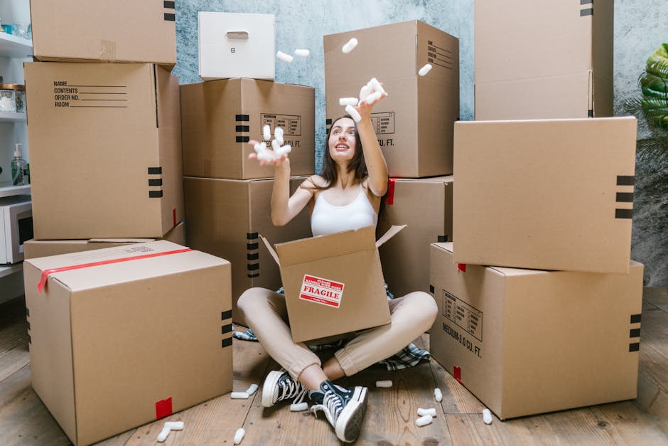 A woman seated on the floor inside a home surrounded by numerous large cardboard moving boxes, some sealed with red packing tape and others open. She is wearing a white sleeveless top and light-colored trousers, smiling as she throws small white packing peanuts into the air above her. The boxes vary in size and are stacked against a light blue wall, with some positioned on the wooden floor and others against the wall. Inside the open box on her lap, there is a red 'Fragile' sticker. To her left, a kitchen area with a white countertop and a small potted plant is partially visible. The scene depicts a home packing and moving process, possibly in preparation for a house relocation, with a focus on packing materials, furniture, and the inside environment prior to professional removal services by Man With a Van Lisson Grove.