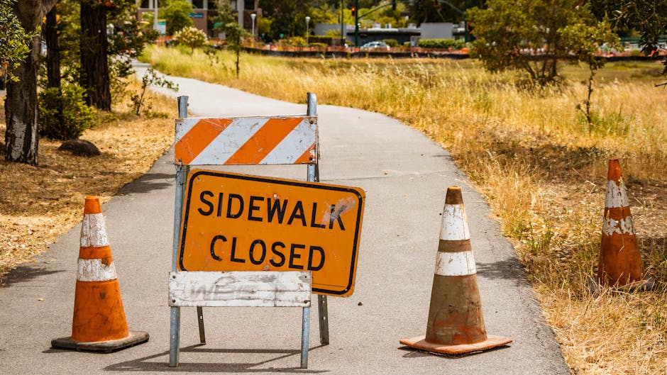 A temporary road closure notice blocks a narrow sidewalk on Lisson Grove, with orange and white traffic cones and a sign reading 'Sidewalk Closed' in black letters on an orange background. The scene depicts a working environment for home relocation or furniture transport, with the sidewalk partially obstructed and a background of trees, grass, and a distant street with parked cars. The sidewalk appears to be in an outdoor urban or suburban area, with a concrete surface and nearby grassy and leafy areas. The traffic management setup facilities the loading or unloading phase of a house removal, managed by Man With a Van Lisson Grove, especially relevant during street closure permits for larger removals in this area. The lighting suggests daytime, with natural sunlight illuminating the scene, emphasizing the temporary nature of the closure involving parking and pedestrian access adjustments for ongoing moving services.