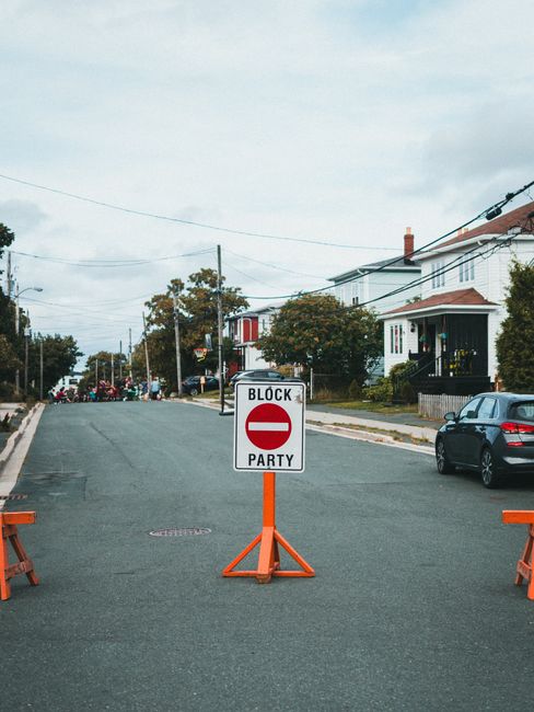 An empty residential street in Lisson Grove with a temporary road closure sign indicating a 'Block Party,' supported by orange barrier stands placed on the pavement. The street features parked cars on the right side, including a black vehicle, with houses and gardens visible in the background under an overcast sky. Utility wires are strung across the road, and trees line the residential area. This setup suggests preparations for a community event or street closure related to local moving or home relocation activities, possibly coordinated by Man With a Van Lisson Grove for logistics involving furniture transport or packing during a house removal.