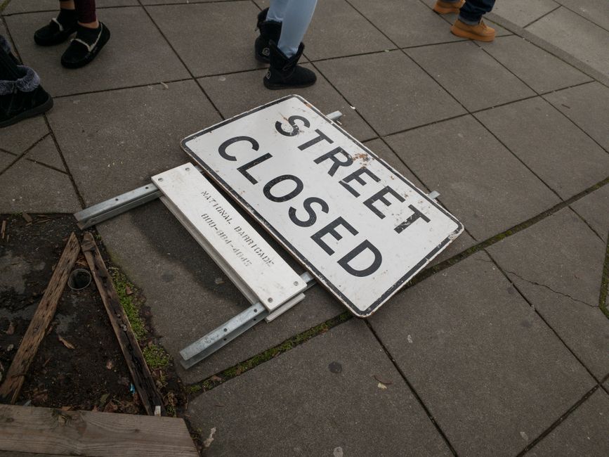 A temporary road closure notice blocks a narrow sidewalk on Lisson Grove, with orange and white traffic cones and a sign reading 'Sidewalk Closed' in black letters on an orange background. The scene depicts a working environment for home relocation or furniture transport, with the sidewalk partially obstructed and a background of trees, grass, and a distant street with parked cars. The sidewalk appears to be in an outdoor urban or suburban area, with a concrete surface and nearby grassy and leafy areas. The traffic management setup facilities the loading or unloading phase of a house removal, managed by Man With a Van Lisson Grove, especially relevant during street closure permits for larger removals in this area. The lighting suggests daytime, with natural sunlight illuminating the scene, emphasizing the temporary nature of the closure involving parking and pedestrian access adjustments for ongoing moving services.